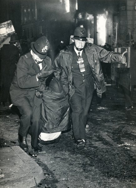 Policemen drag away an injured colleague as buildings burn during the Toxteth Riots